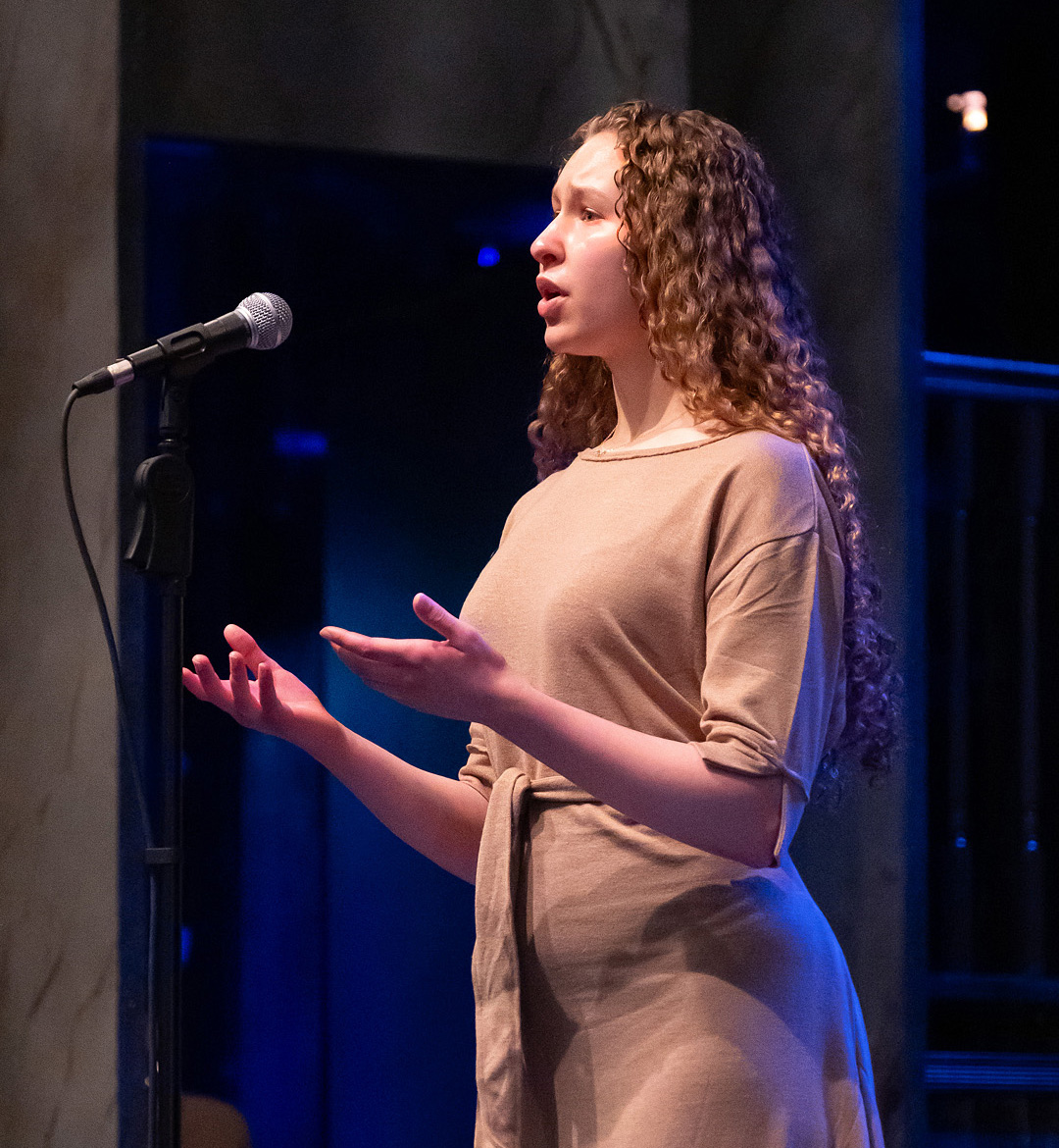 Ellette Whitcomb, with long curly blond hair and wearing beige dress, gestures with her hands as she recites