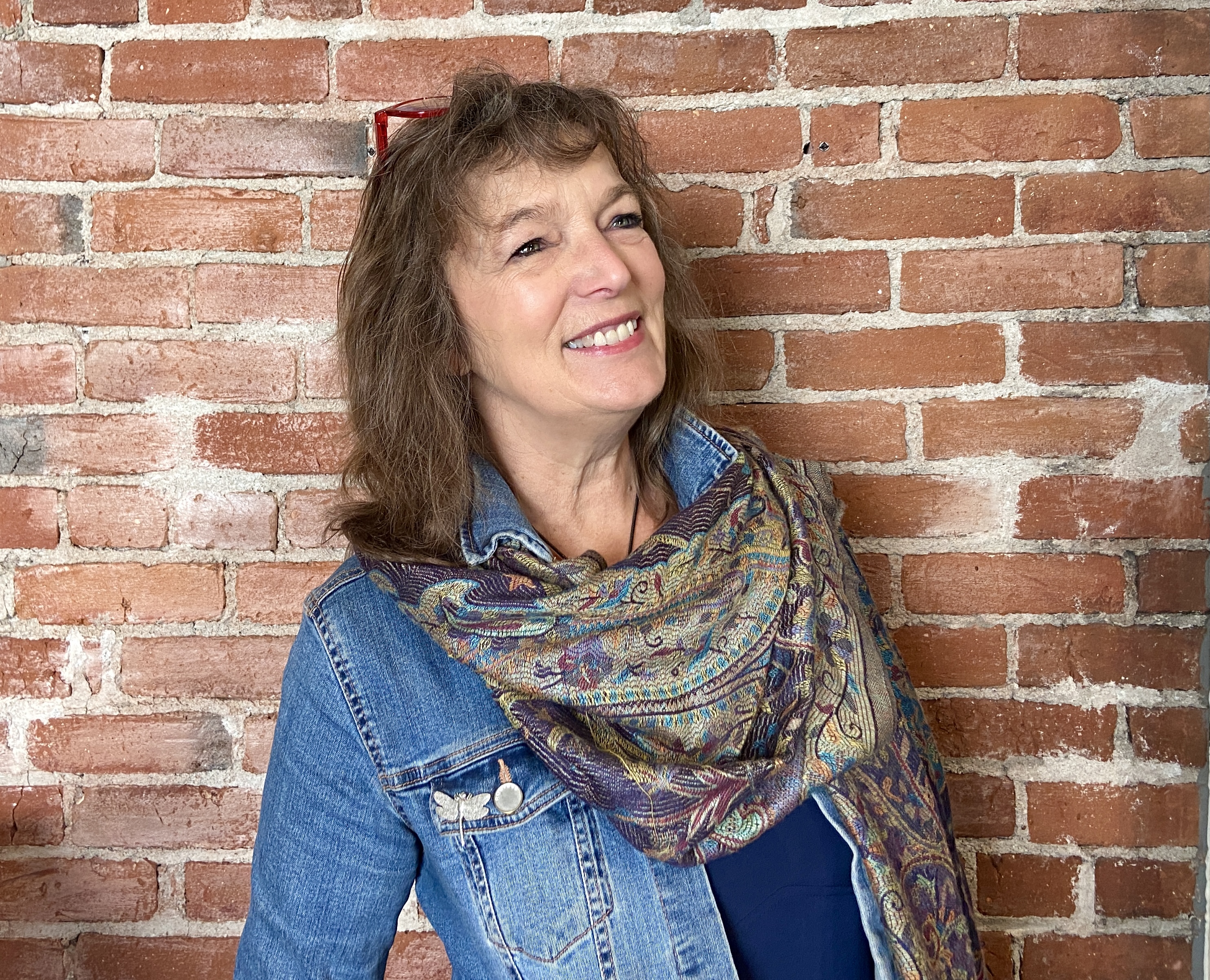 Portrait of Krys Holmes, wearing denim jacket and multicolored scarf, smiling and standing in front of brick wall.